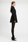 A model in profile wearing a black long-sleeve mini dress with a tie waist, black tights, and black pointed-toe heels, photographed against a plain light gray studio background.