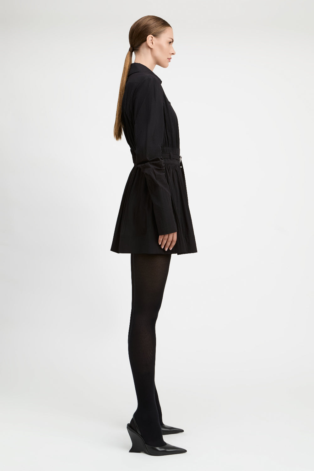 A model in profile wearing a black long-sleeve mini dress with a tie waist, black tights, and black pointed-toe heels, photographed against a plain light gray studio background.