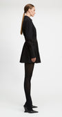 A model in profile wearing a black long-sleeve mini dress with a tie waist, black tights, and black pointed-toe heels, photographed against a plain light gray studio background.