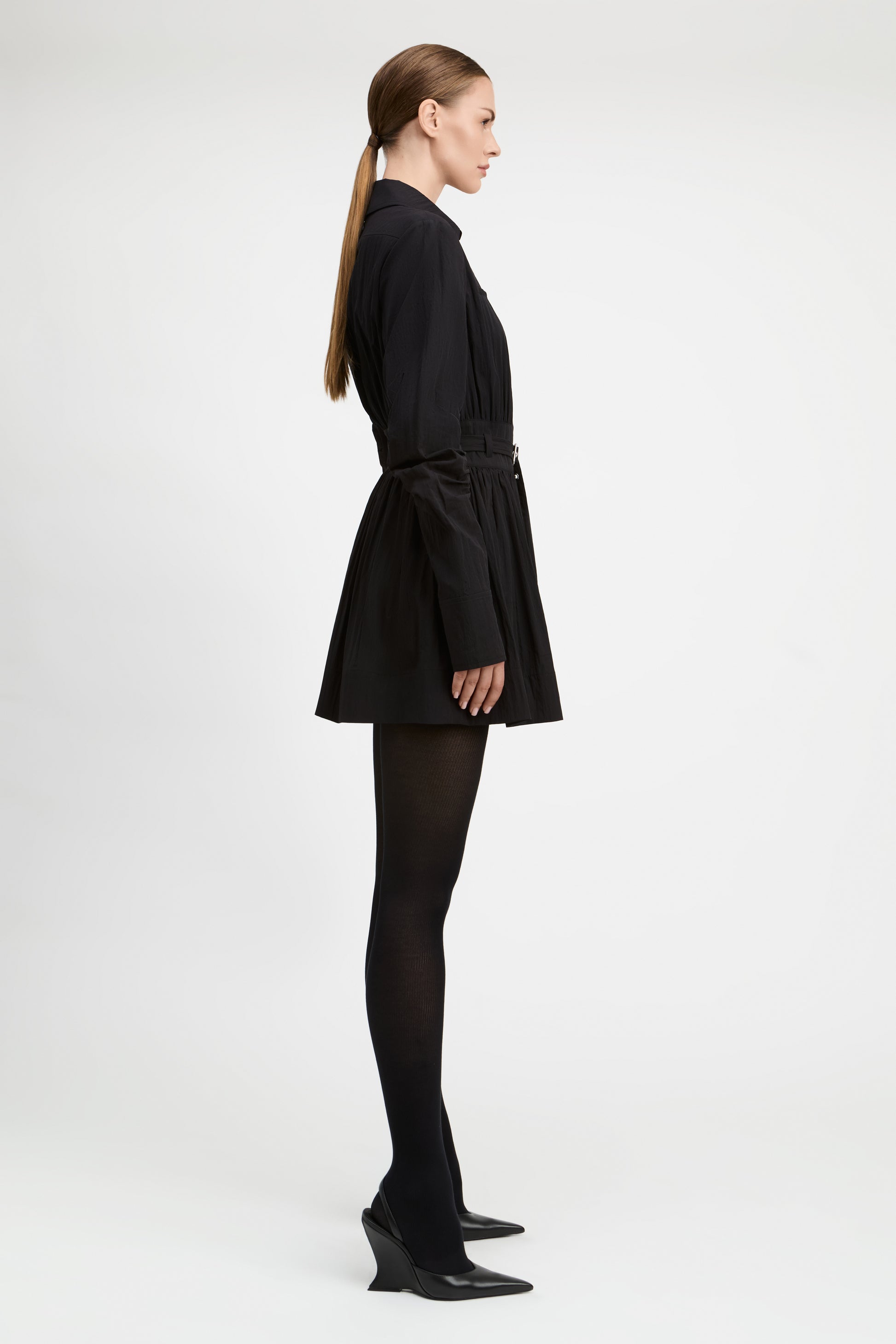 A model in profile wearing a black long-sleeve mini dress with a tie waist, black tights, and black pointed-toe heels, photographed against a plain light gray studio background.