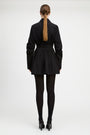 A model viewed from the back wearing a black long-sleeve mini jacket with a pleated skirt and waist belt, paired with black tights and black heels, against a light gray studio background.