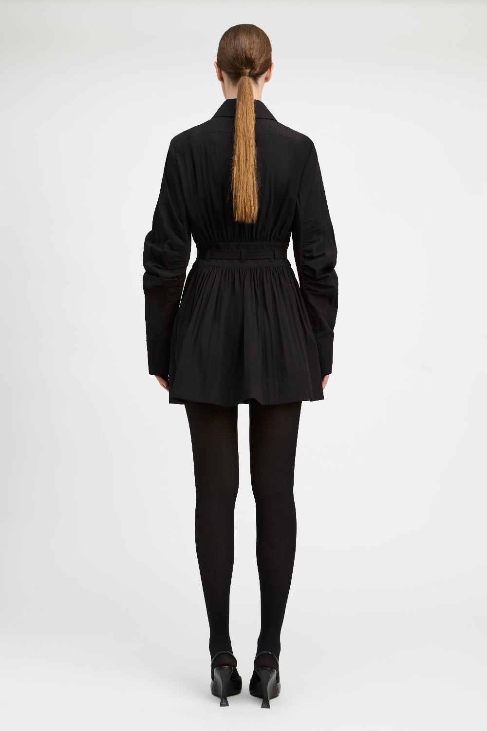 A model viewed from the back wearing a black long-sleeve mini jacket with a pleated skirt and waist belt, paired with black tights and black heels, against a light gray studio background.
