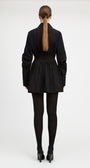 A model viewed from the back wearing a black long-sleeve mini jacket with a pleated skirt and waist belt, paired with black tights and black heels, against a light gray studio background.