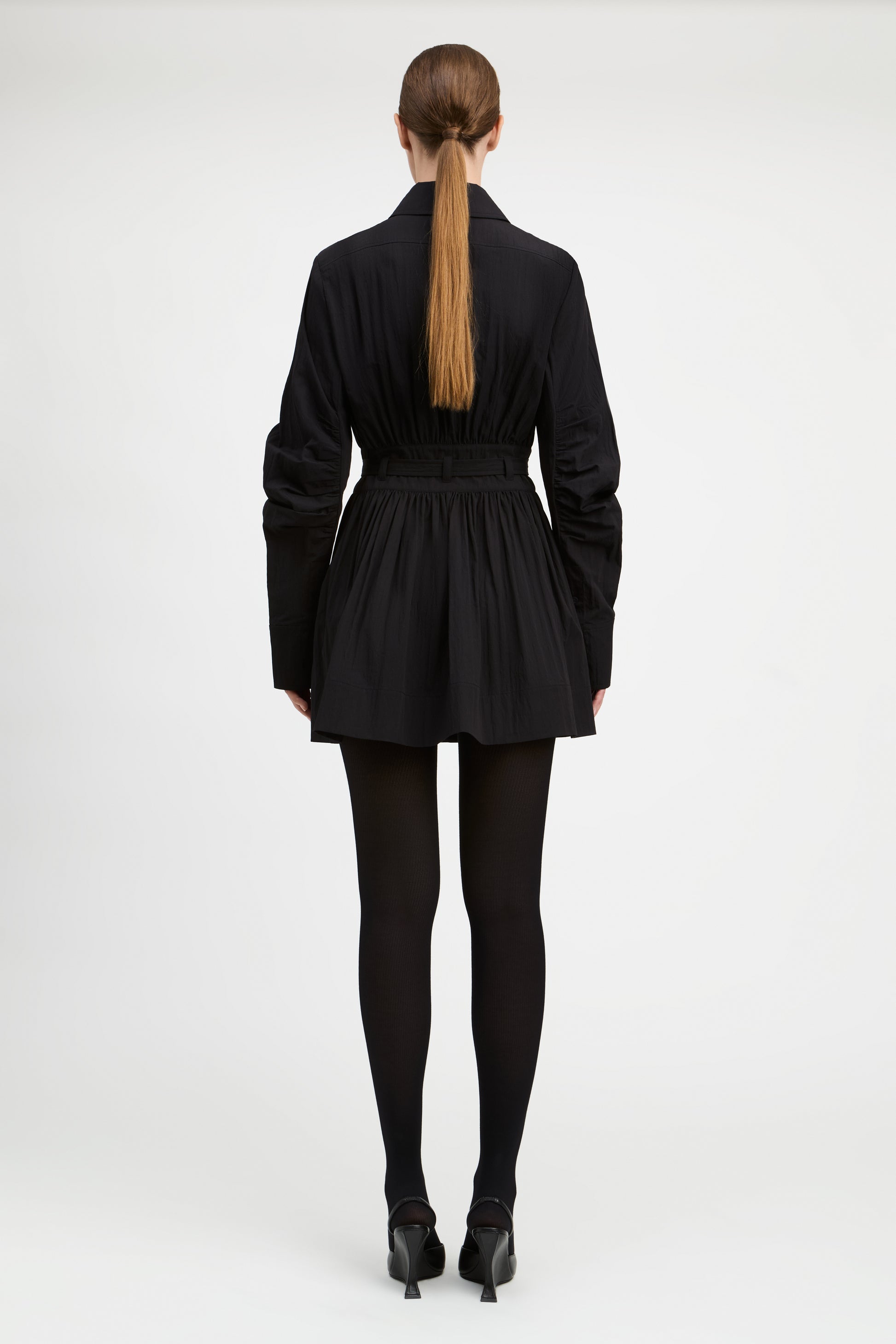 A model viewed from the back wearing a black long-sleeve mini jacket with a pleated skirt and waist belt, paired with black tights and black heels, against a light gray studio background.