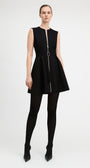 A model wearing a sleeveless black mini dress with a front zipper, a matching black belt with a silver buckle, black tights, and black pointed-toe heels, photographed against a light gray studio background.