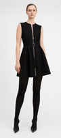 A model wearing a sleeveless black mini dress with a front zipper, a matching black belt with a silver buckle, black tights, and black pointed-toe heels, photographed against a light gray studio background.