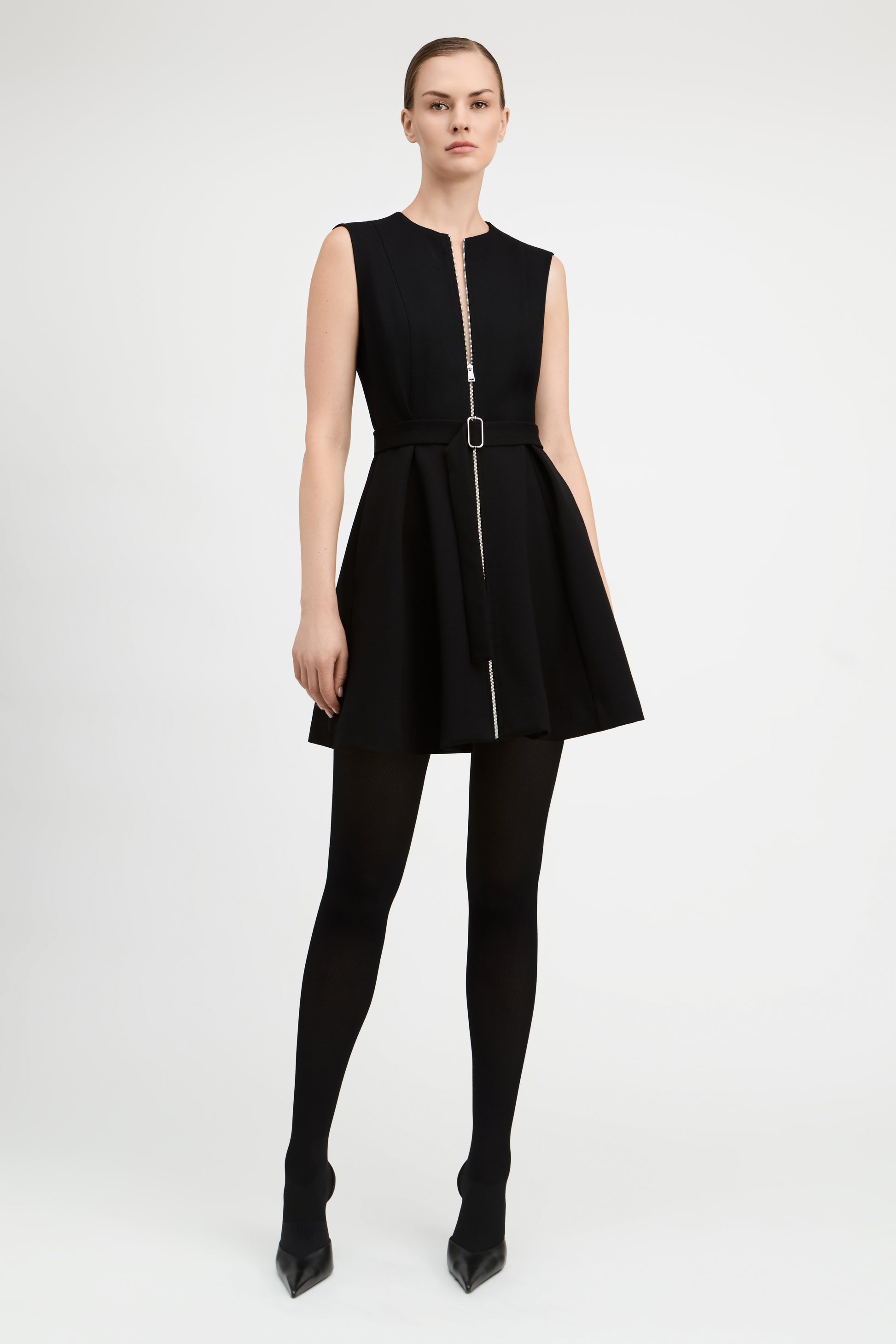 A model wearing a sleeveless black mini dress with a front zipper, a matching black belt with a silver buckle, black tights, and black pointed-toe heels, photographed against a light gray studio background.