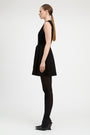 A woman in a side profile wearing a sleeveless black mini dress with a tie waist, black tights, and black wedge heels, photographed against a plain light gray studio background.