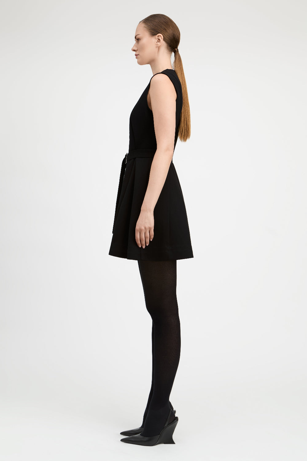 A woman in a side profile wearing a sleeveless black mini dress with a tie waist, black tights, and black wedge heels, photographed against a plain light gray studio background.