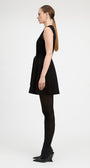 A woman in a side profile wearing a sleeveless black mini dress with a tie waist, black tights, and black wedge heels, photographed against a plain light gray studio background.