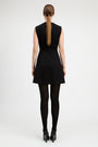A woman viewed from the back wearing a sleeveless black mini dress with a matching waist tie, black tights, and black heels against a light gray studio background.