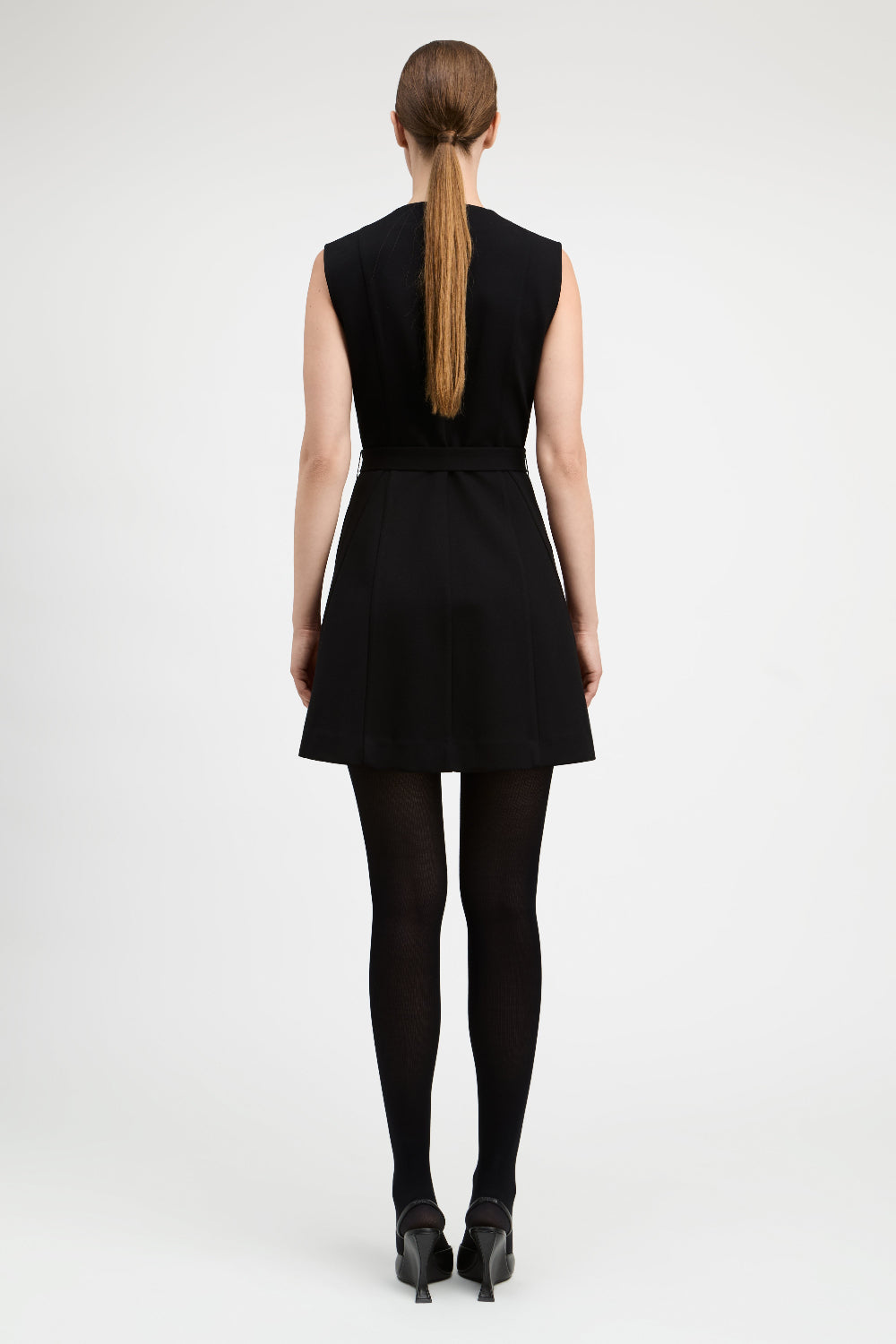 A woman viewed from the back wearing a sleeveless black mini dress with a matching waist tie, black tights, and black heels against a light gray studio background.