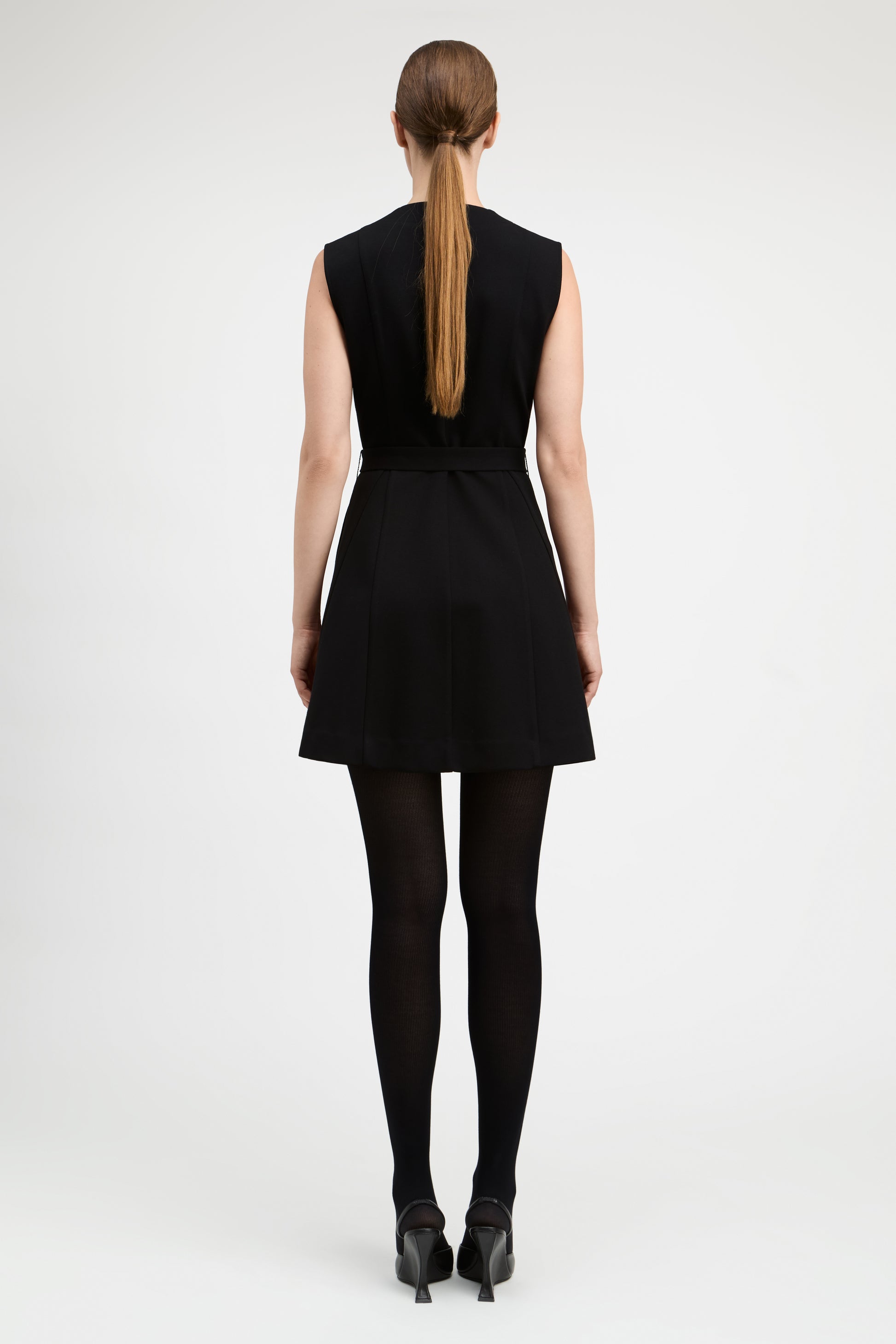 A woman viewed from the back wearing a sleeveless black mini dress with a matching waist tie, black tights, and black heels against a light gray studio background.