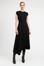 A model wearing a black high-low midi dress with cap sleeves and black leather ankle boots, standing against a light gray studio background.