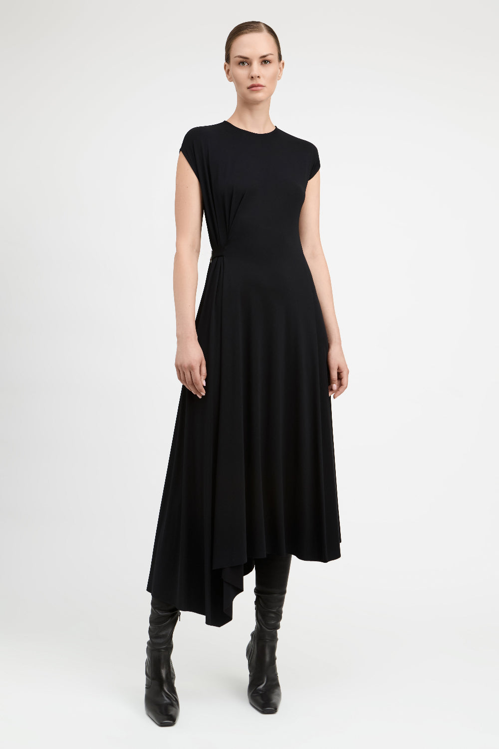 A model wearing a black high-low midi dress with cap sleeves and black leather ankle boots, standing against a light gray studio background.