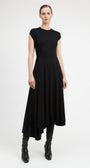 A model wearing a black high-low midi dress with cap sleeves and black leather ankle boots, standing against a light gray studio background.
