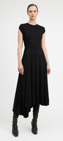 A model wearing a black high-low midi dress with cap sleeves and black leather ankle boots, standing against a light gray studio background.