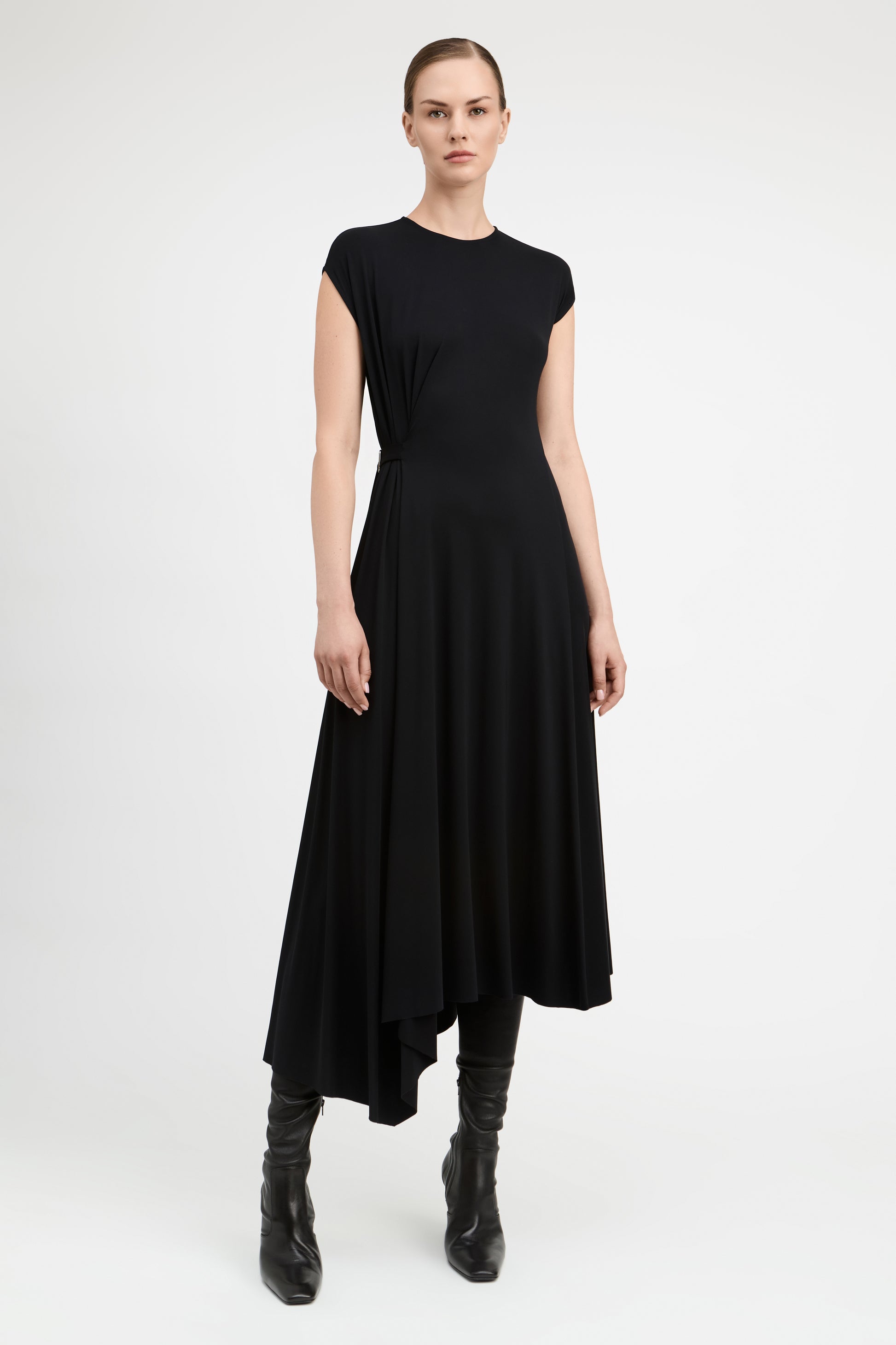 A model wearing a black high-low midi dress with cap sleeves and black leather ankle boots, standing against a light gray studio background.