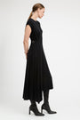 A woman modeling a black midi dress with a belted waist, shown from a side profile against a plain light gray studio background.
