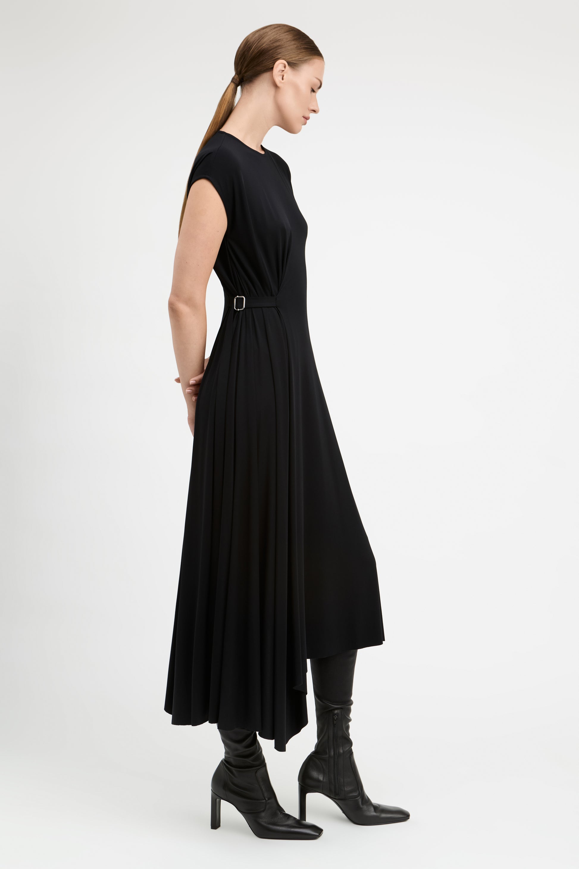 A woman modeling a black midi dress with a belted waist, shown from a side profile against a plain light gray studio background.