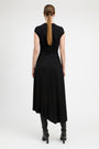 A woman viewed from the back wearing a black sleeveless midi dress with a gathered waist and black heeled ankle boots, against a plain light gray studio background.