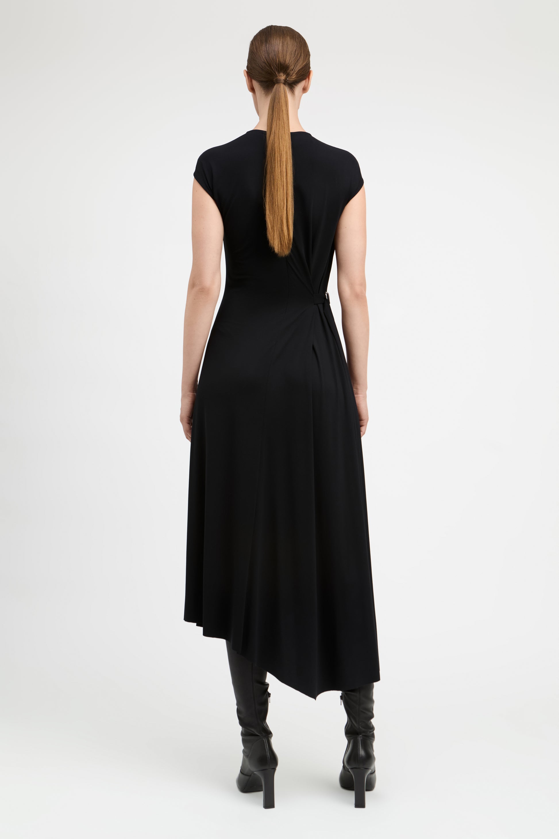 A woman viewed from the back wearing a black sleeveless midi dress with a gathered waist and black heeled ankle boots, against a plain light gray studio background.
