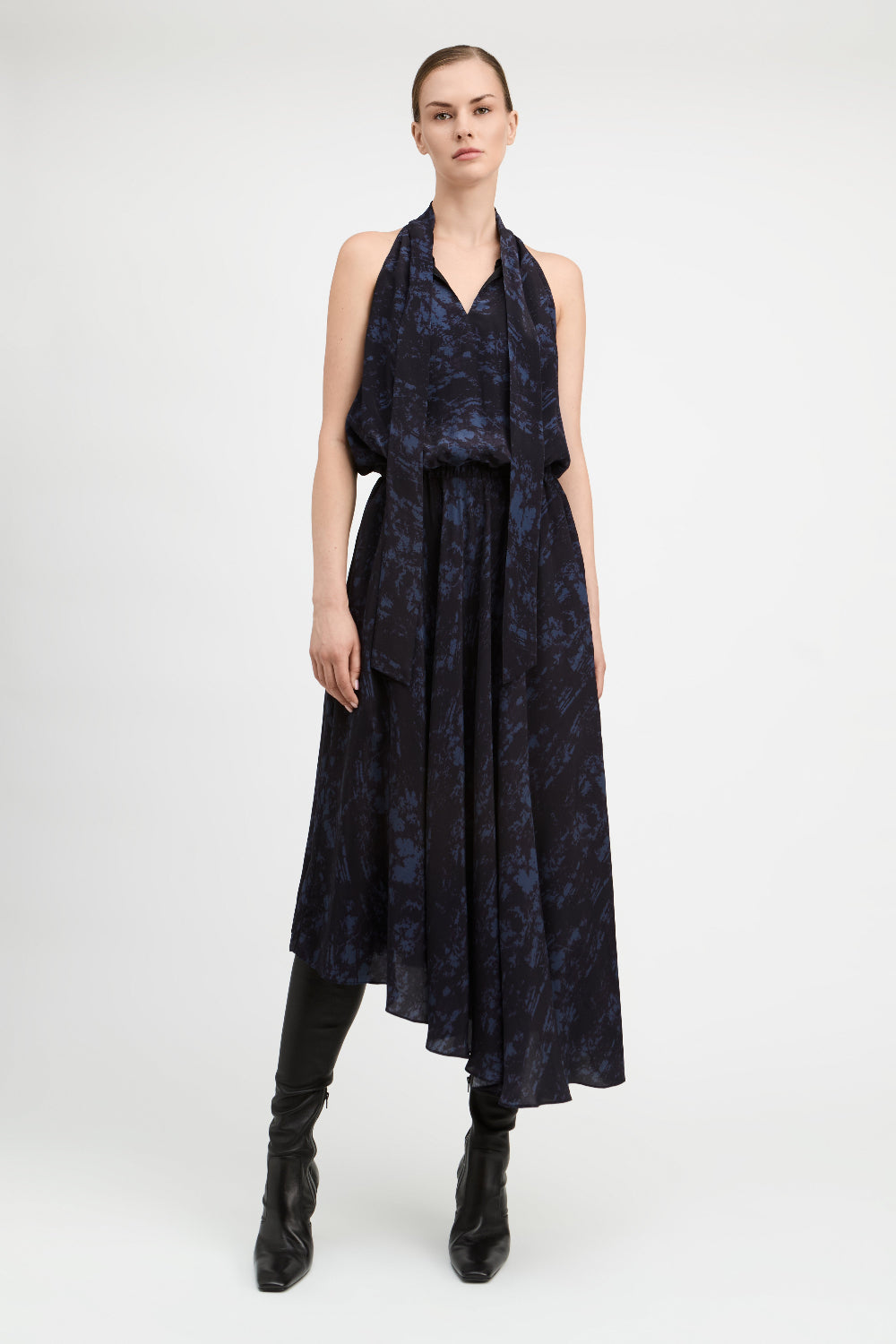 A model stands against a light gray studio background wearing a sleeveless dark navy midi dress with a blue abstract pattern, black knee-high boots, and black leggings.