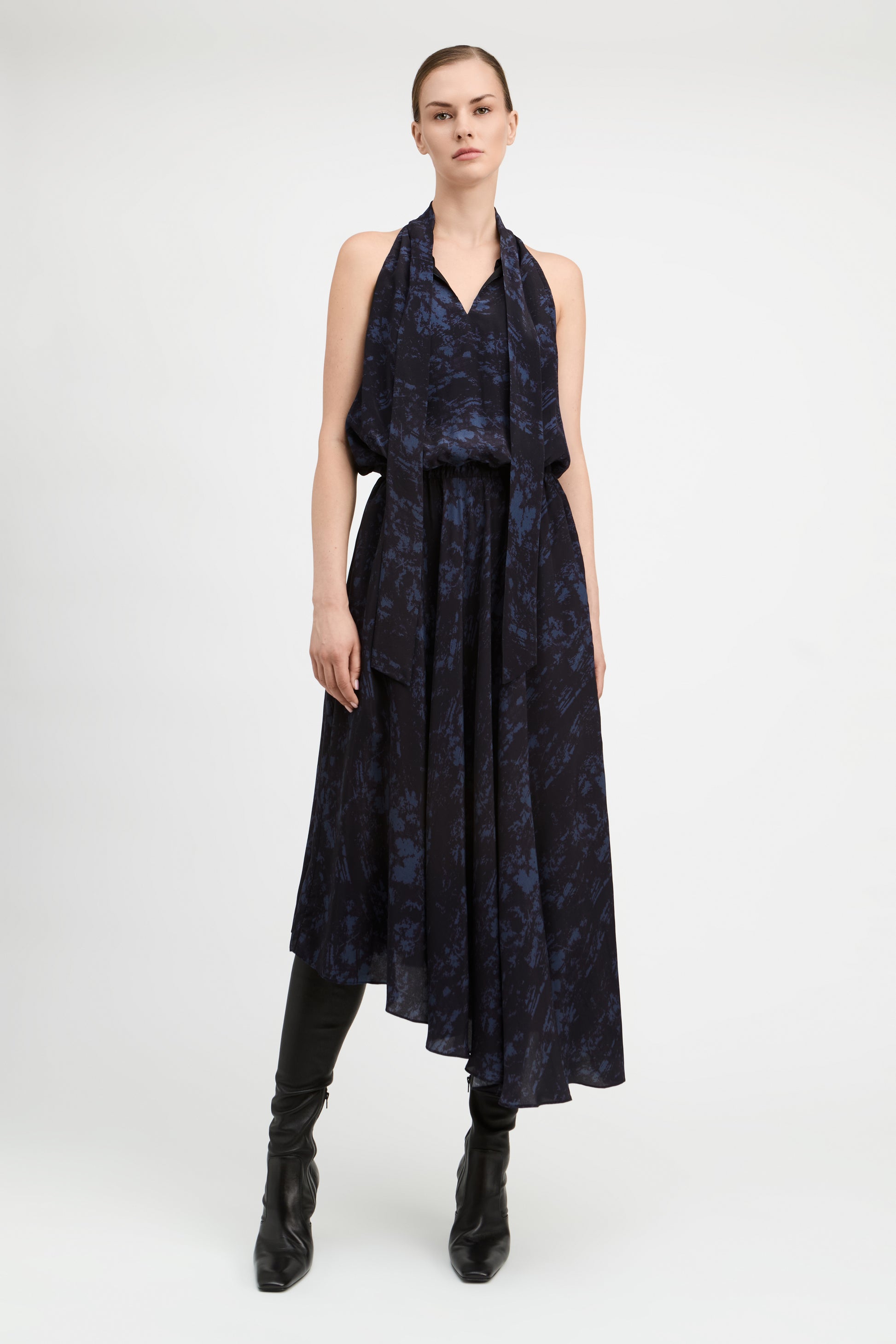 A model stands against a light gray studio background wearing a sleeveless dark navy midi dress with a blue abstract pattern, black knee-high boots, and black leggings.