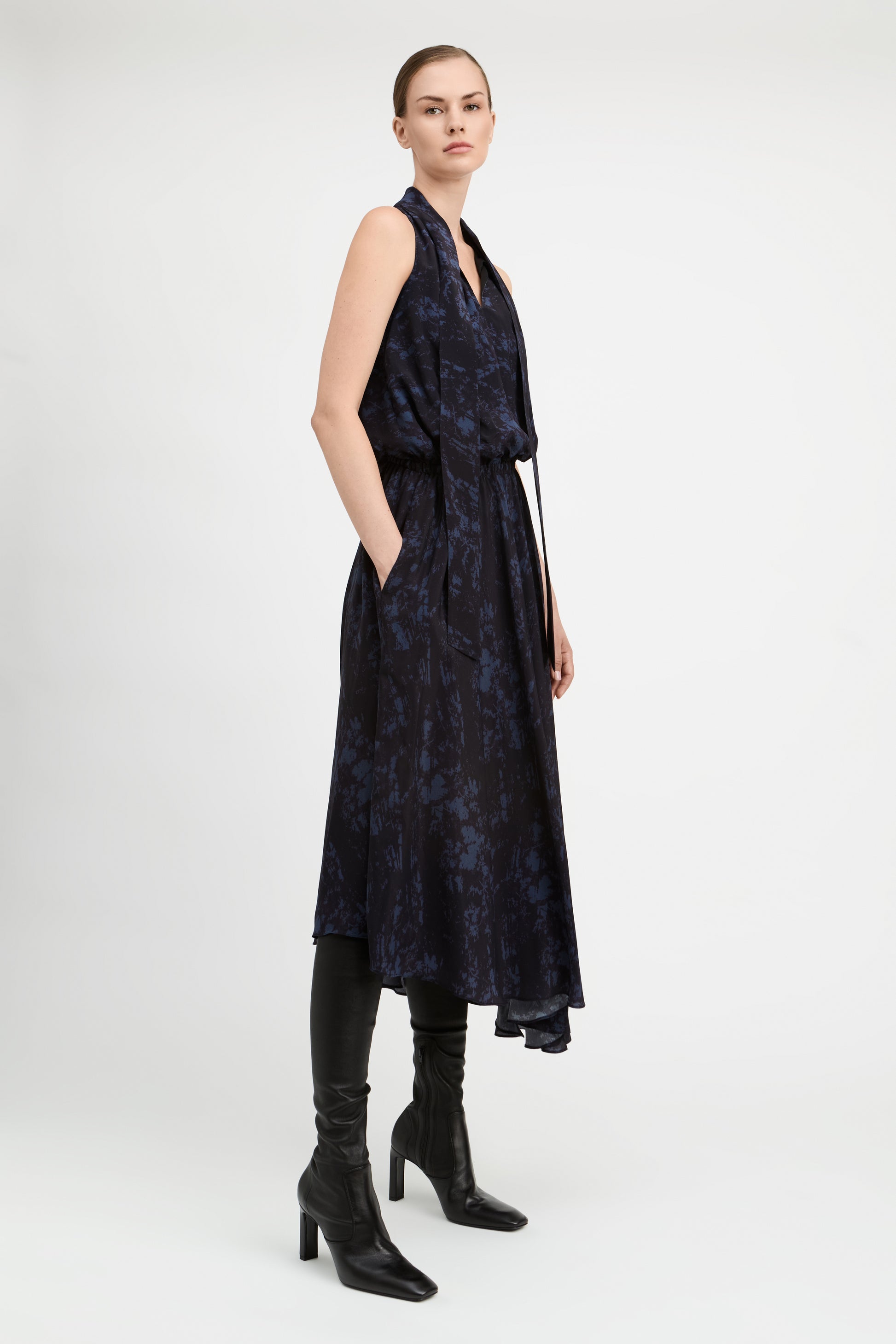 A model wearing a sleeveless dark navy midi dress with a subtle tonal floral pattern, styled with black knee-high leather boots against a light gray studio background.