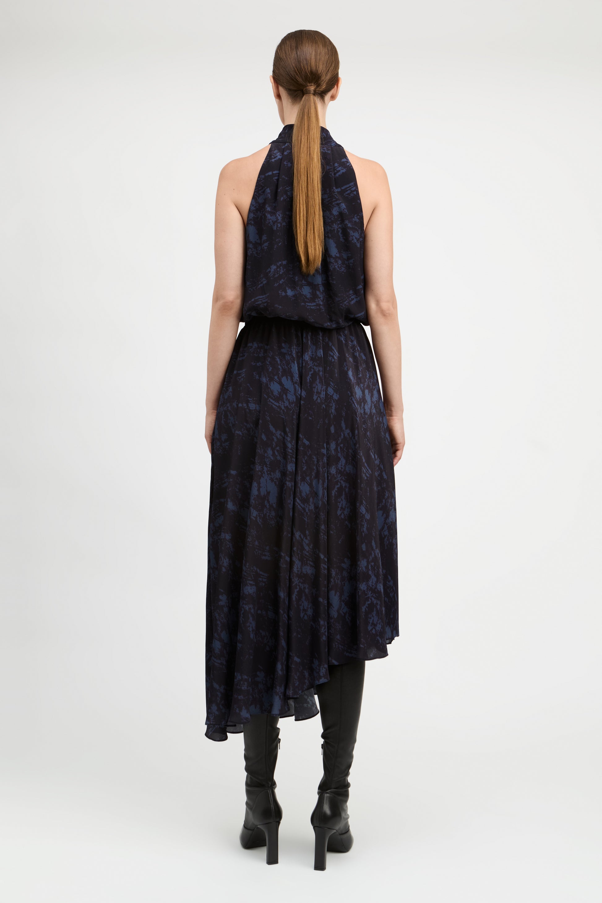 A woman viewed from the back wearing a sleeveless dark navy dress with a blue abstract print and black knee-high boots against a light gray studio background.