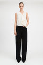 A model stands against a light gray studio background wearing a sleeveless cream wrap-style top with draped fabric and black wide-leg trousers.