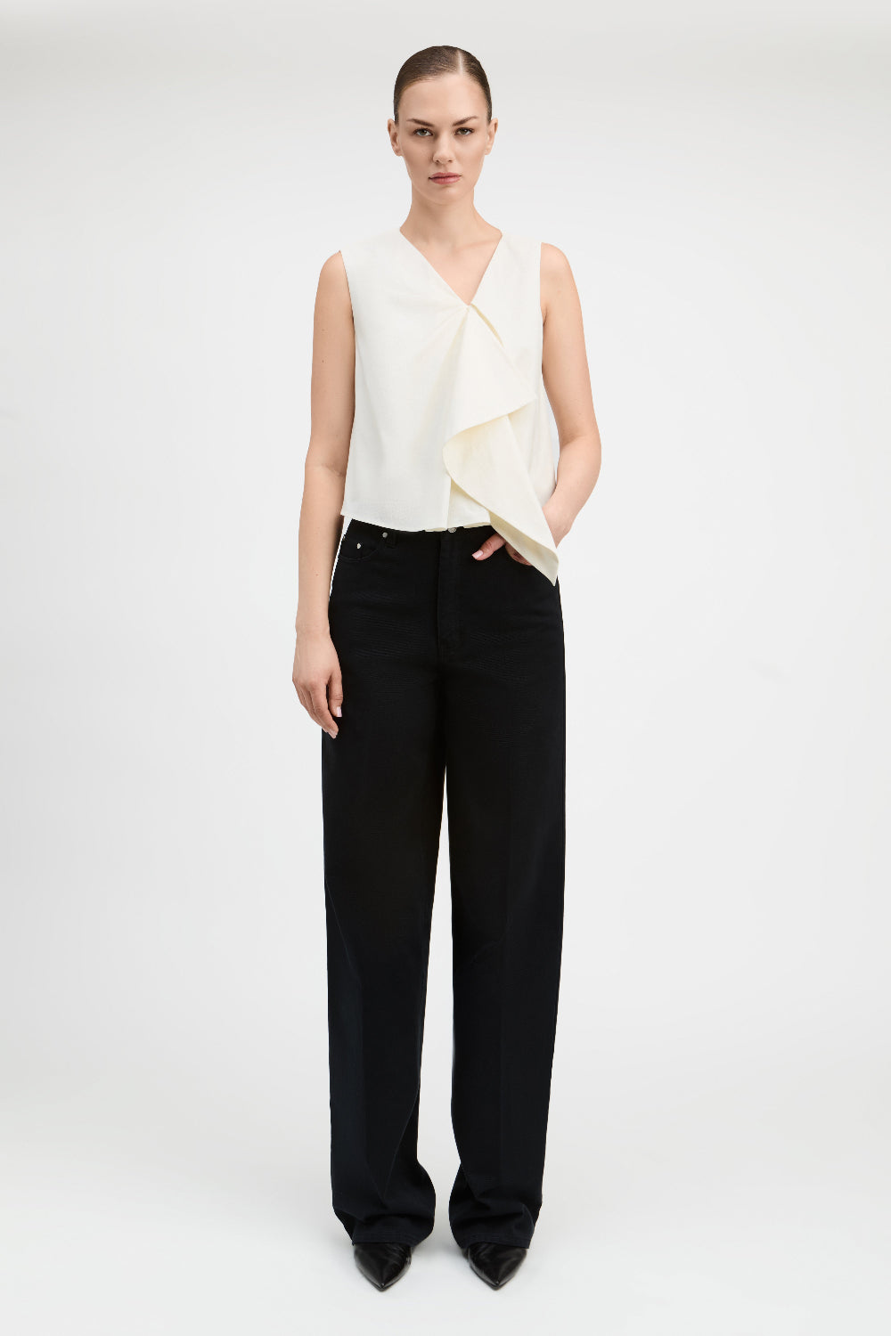 A model stands against a light gray studio background wearing a sleeveless cream wrap-style top with draped fabric and black wide-leg trousers.