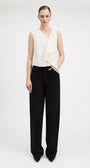 A model stands against a light gray studio background wearing a sleeveless cream wrap-style top with draped fabric and black wide-leg trousers.