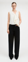 A model stands against a light gray studio background wearing a sleeveless cream wrap-style top with draped fabric and black wide-leg trousers.