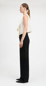 A woman stands in profile wearing a sleeveless cream draped top and wide-leg black trousers against a plain light gray studio background.
