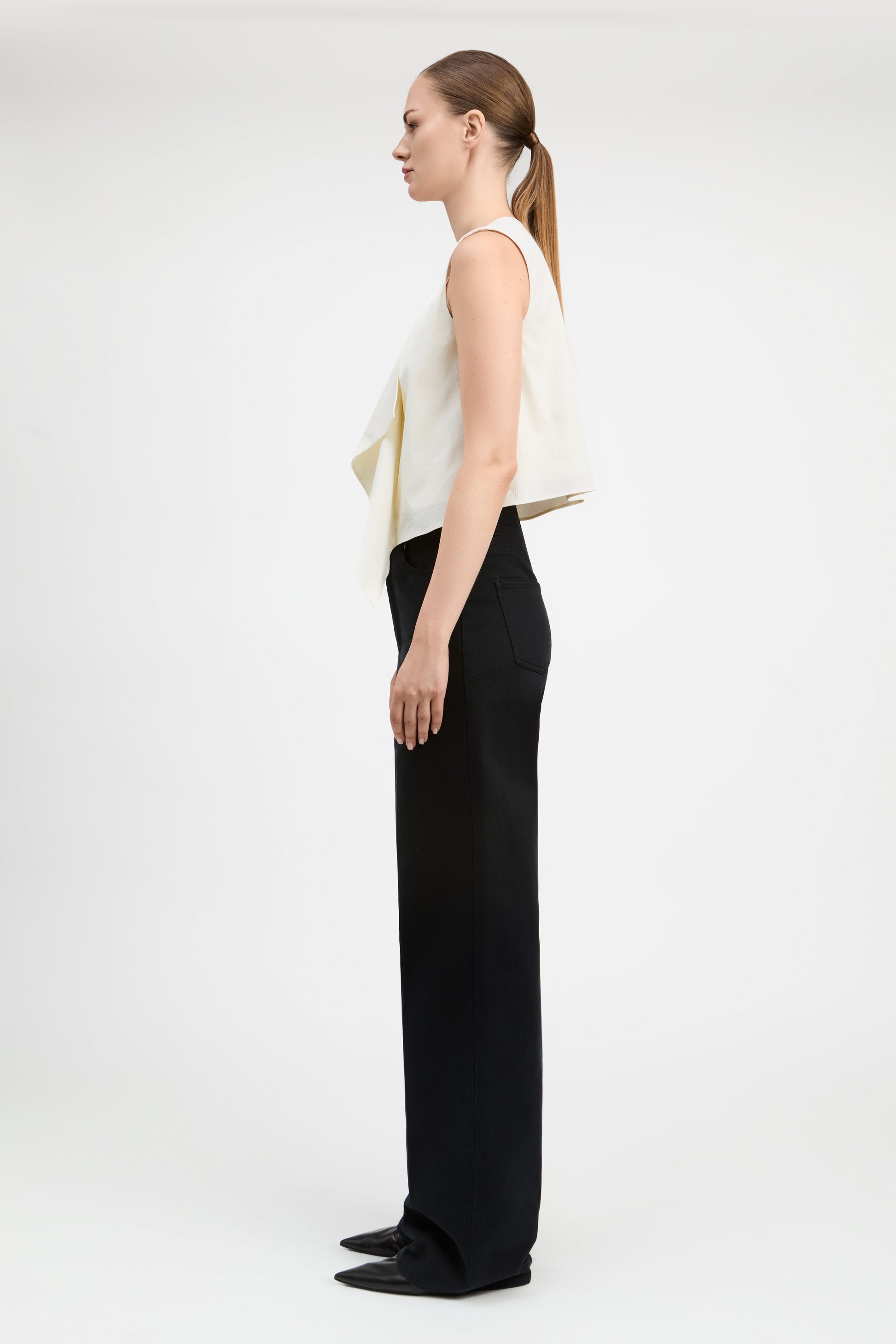 A woman stands in profile wearing a sleeveless cream draped top and wide-leg black trousers against a plain light gray studio background.
