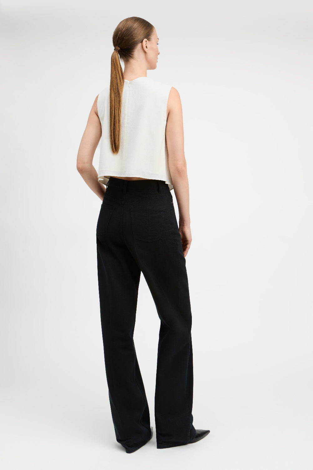 A woman shown from the back wearing a sleeveless white top and black wide-leg trousers against a plain light gray studio background.