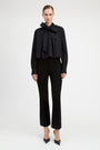 A model wearing a black long-sleeve blouse with a dramatic ruffled bow collar and matching black cropped trousers, standing against a plain light gray studio background.