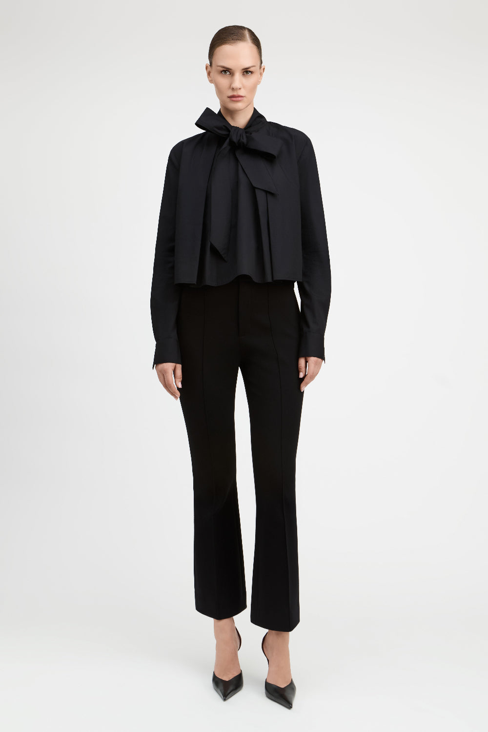 A model wearing a black long-sleeve blouse with a dramatic ruffled bow collar and matching black cropped trousers, standing against a plain light gray studio background.