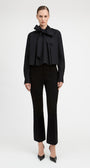 A model wearing a black long-sleeve blouse with a dramatic ruffled bow collar and matching black cropped trousers, standing against a plain light gray studio background.