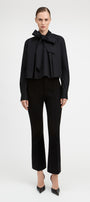 A model wearing a black long-sleeve blouse with a dramatic ruffled bow collar and matching black cropped trousers, standing against a plain light gray studio background.