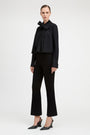 A model stands against a light gray studio background wearing a black long-sleeve blouse with a large bow at the collar, black cropped flared pants, and black pointed slingback heels.