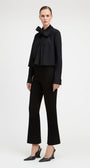 A model stands against a light gray studio background wearing a black long-sleeve blouse with a large bow at the collar, black cropped flared pants, and black pointed slingback heels.