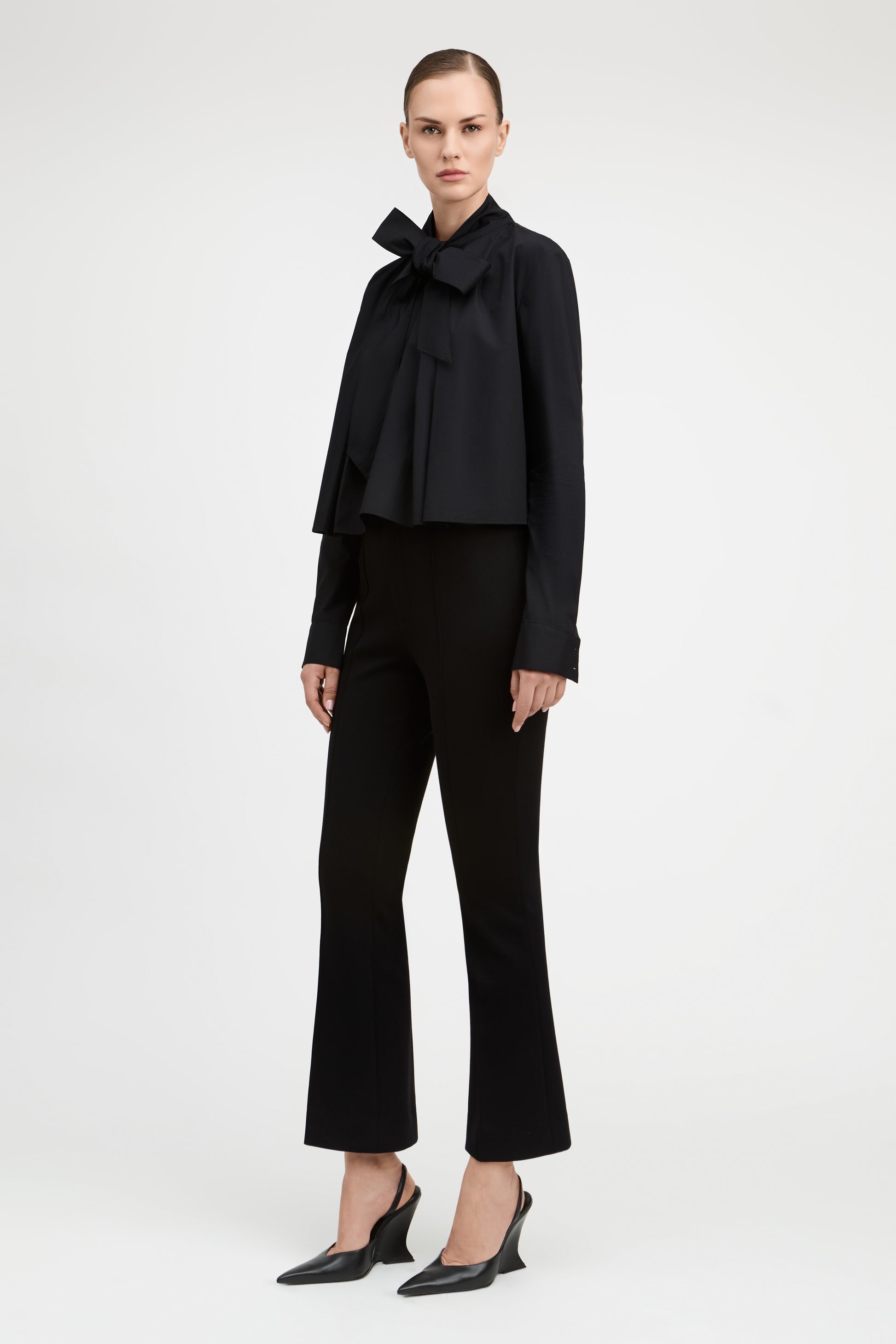 A model stands against a light gray studio background wearing a black long-sleeve blouse with a large bow at the collar, black cropped flared pants, and black pointed slingback heels.