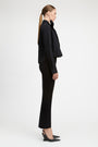 A model in side profile wearing a black long-sleeve cropped jacket with a large bow collar, black flared trousers, and black pointed-toe heels against a light gray studio background.