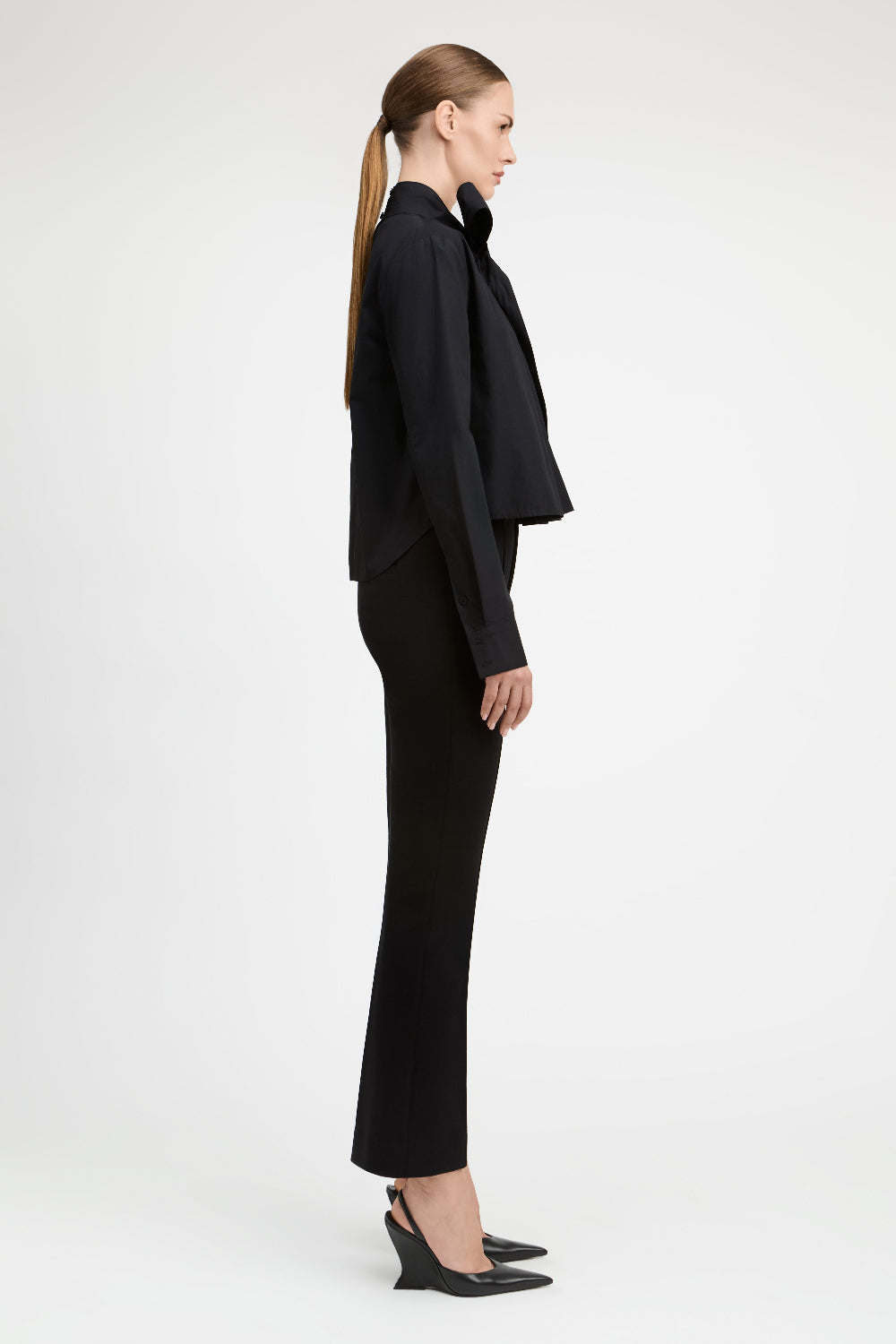 A model in side profile wearing a black long-sleeve cropped jacket with a large bow collar, black flared trousers, and black pointed-toe heels against a light gray studio background.