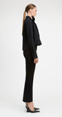 A model in side profile wearing a black long-sleeve cropped jacket with a large bow collar, black flared trousers, and black pointed-toe heels against a light gray studio background.
