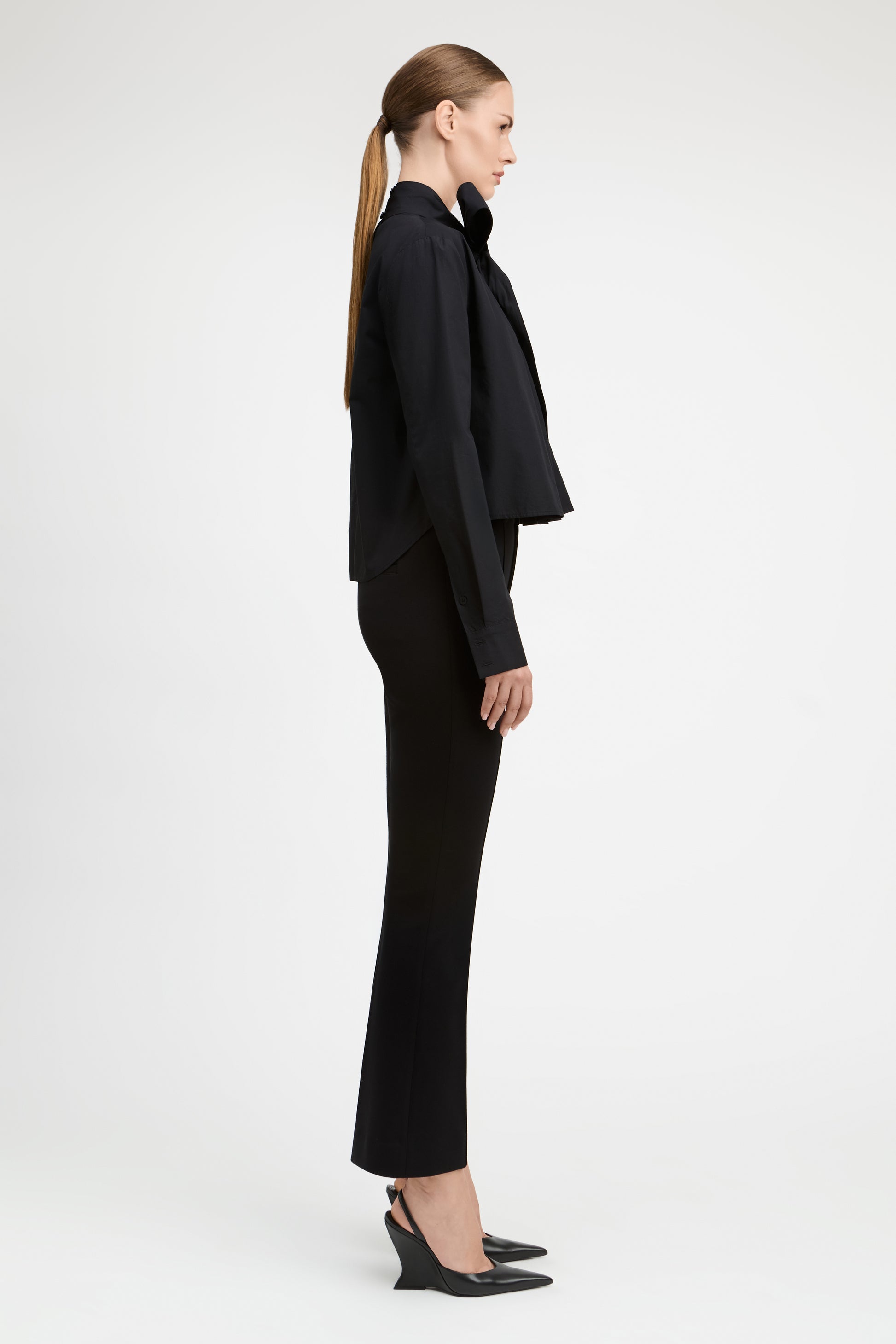 A model in side profile wearing a black long-sleeve cropped jacket with a large bow collar, black flared trousers, and black pointed-toe heels against a light gray studio background.