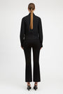 A woman viewed from the back wearing a black long-sleeve mock-neck top and matching black flared pants, paired with black heels, against a plain light gray studio background.