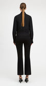 A woman viewed from the back wearing a black long-sleeve mock-neck top and matching black flared pants, paired with black heels, against a plain light gray studio background.